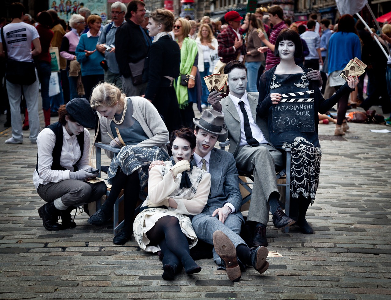 Six street performers wearing white mime makeup and clothes in various shades of black, white, and gray pose in a tableau. They are sitting in pairs and appearing to have conversations with each other. Behind them a large crowd of passers-by mills.